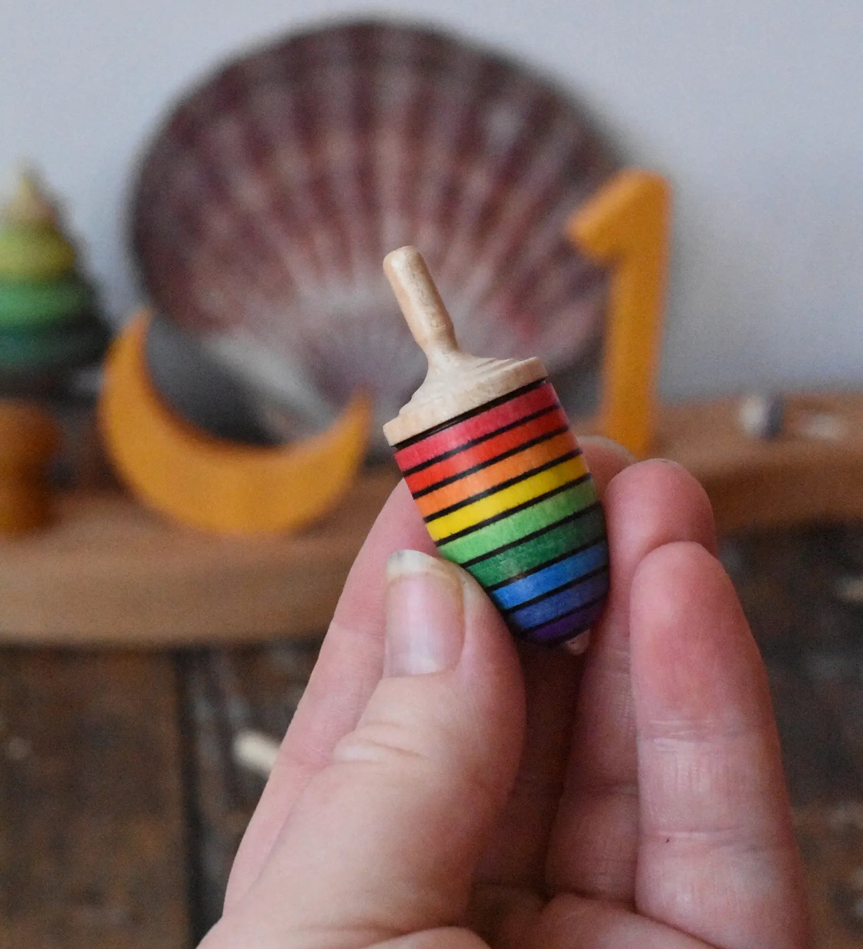 Mader handcrafted wooden rainbow mini thunderbolt spinning top in an adult's hand with a decorated Grimm's celebration ring in the background.