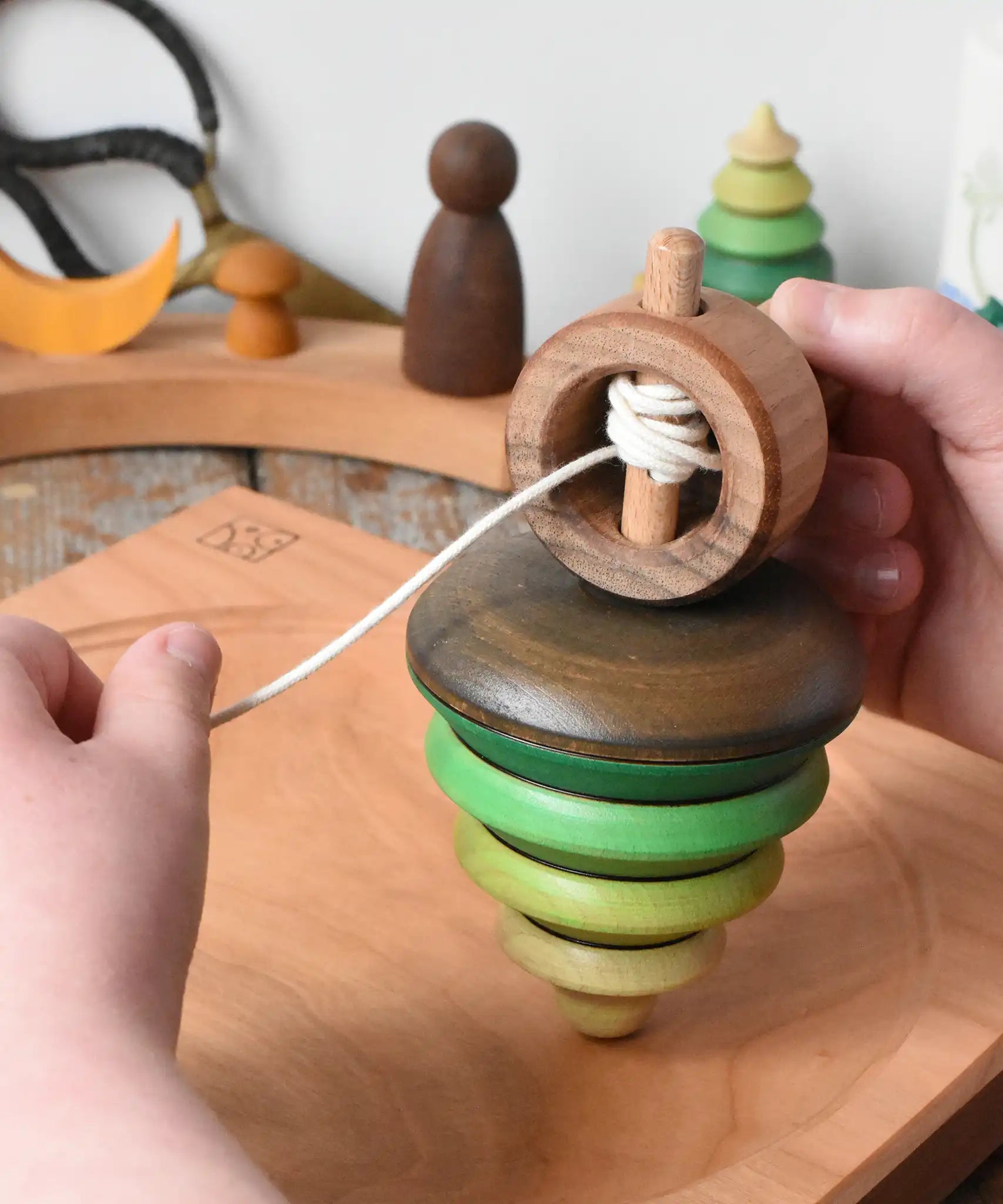 Mader pull-string tree spinning top placed on a cherry wood spinning plate in the Babipur playroom. A child'd hand can be seen holding the wooden handle and pulling the string.