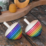 Two Mader Kreiselmanufaktur rainbow egg spinning top placed on a wooden surface in the Babipur playroom. Both have rainbow stripes, one starting with the red colour the other with purple.