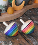 Two Mader Kreiselmanufaktur rainbow egg spinning top placed on a wooden surface in the Babipur playroom. Both have rainbow stripes, one starting with the red colour the other with purple.