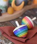 Mader Kreiselmanufaktur rainbow egg spinning top placed on a cloth on a wooden surface in the Babipur playroom. Another spinner can be seen in the background. 
