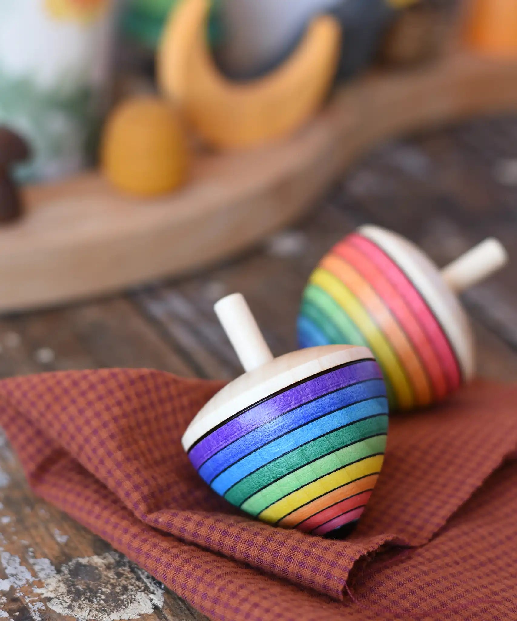 Mader Kreiselmanufaktur rainbow egg spinning top placed on a cloth on a wooden surface in the Babipur playroom. Another spinner can be seen in the background. 