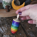 Mader rainbow stripe wooden spinning top placed on a wooden desk in the Babipur playroom. An adult's hand is holding the spinner upright.
