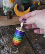 Mader rainbow stripe wooden spinning top placed on a wooden desk in the Babipur playroom. An adult's hand is holding the spinner upright.