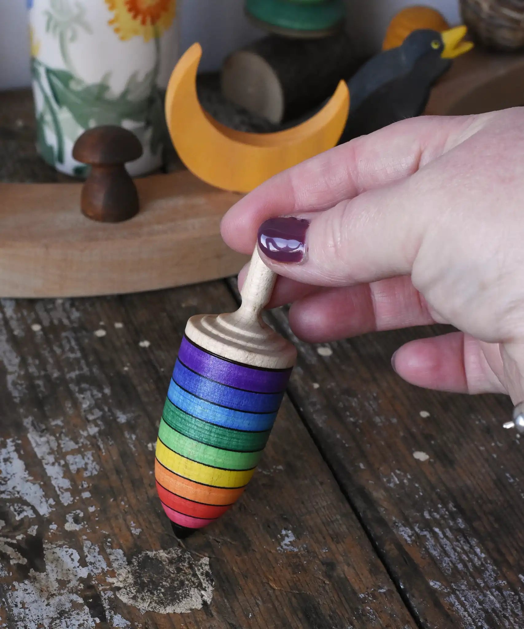 Mader rainbow stripe wooden spinning top placed on a wooden desk in the Babipur playroom. An adult's hand is holding the spinner upright.