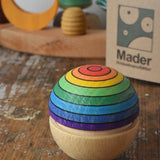 Mader Kreiselmanufaktur rainbow wobble placed on a wooden desk in the Babipur playroom. The cardboard packaging box can be seen in the background along with a decorated Grimm's celebration ring. 