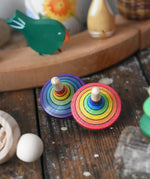 Two rainbow rallye style traditional striped rainbow spinning top toys placed on a wooden desk in the Babipur playroom.