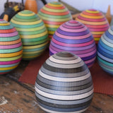 Mader Kreiselmanufaktur roly-poly wooden egg in graphite placed on a wooden surface in the Babipur playroom. Other colour ways of the egg can be seen in the background. 