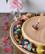 Mader Kreiselmanufaktur 40cm Rondell Ash Wood Spinning Plate placed on a wooden surface in the Babipur playroom. Monalotte spinning top placed on the top with other spinning tops filling bottom part of the plate. 