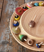 Mader Kreiselmanufaktur 40cm Rondell Ash Wood Spinning Plate placed on a wooden surface in the Babipur playroom. Dice spinning top placed on the top with other spinning tops filling bottom part of the plate. 