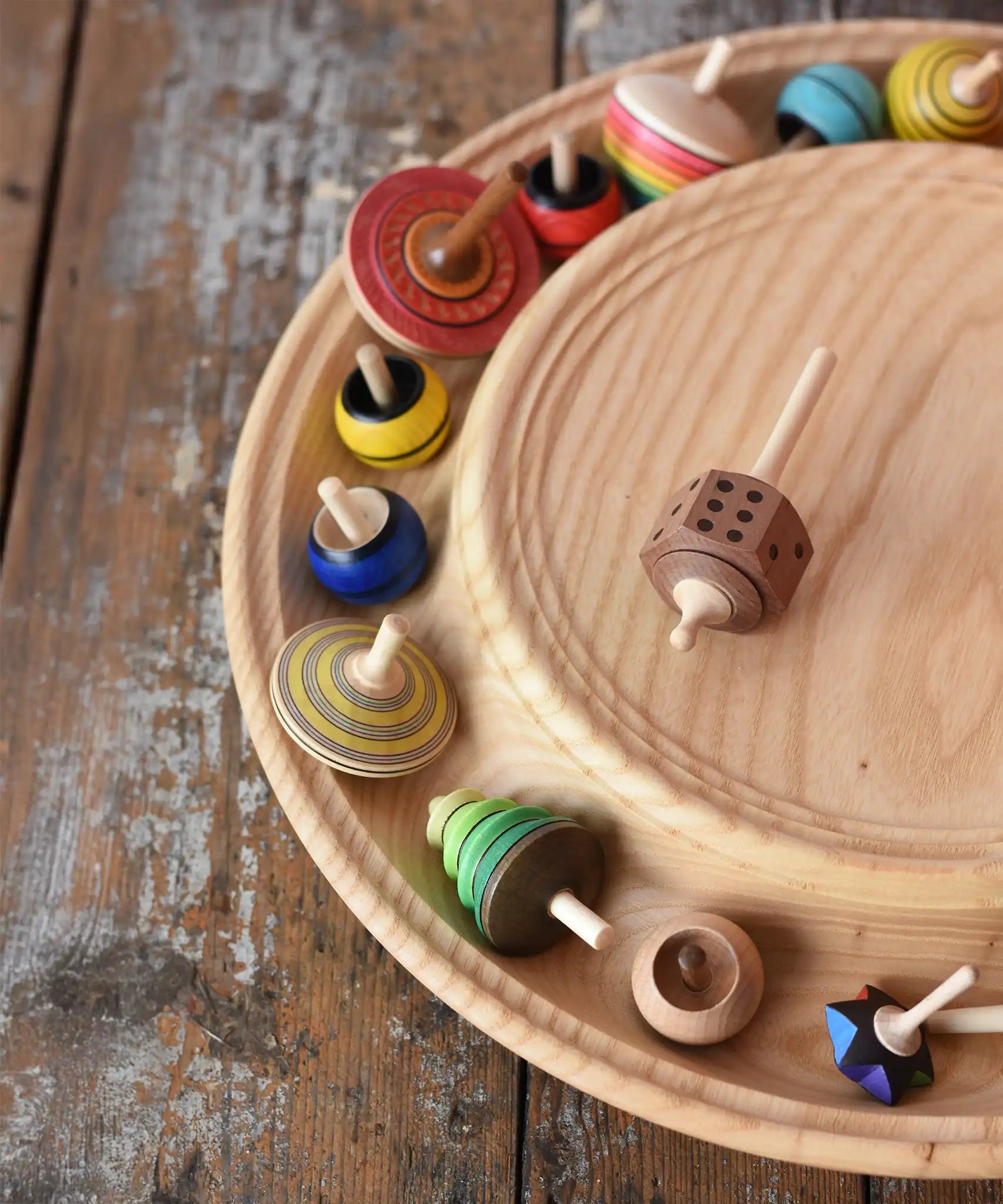 Mader Kreiselmanufaktur 40cm Rondell Ash Wood Spinning Plate placed on a wooden surface in the Babipur playroom. Dice spinning top placed on the top with other spinning tops filling bottom part of the plate. 