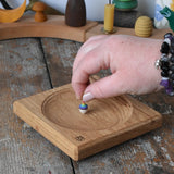 Mader Small Oak Spinning Plate placed on a wooden surface in the Babipur playroom. An adult's hand is placing a miniature rainbow spinning top on the board.