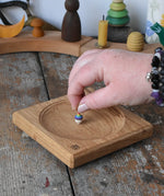 Mader Small Oak Spinning Plate placed on a wooden surface in the Babipur playroom. An adult's hand is placing a miniature rainbow spinning top on the board.