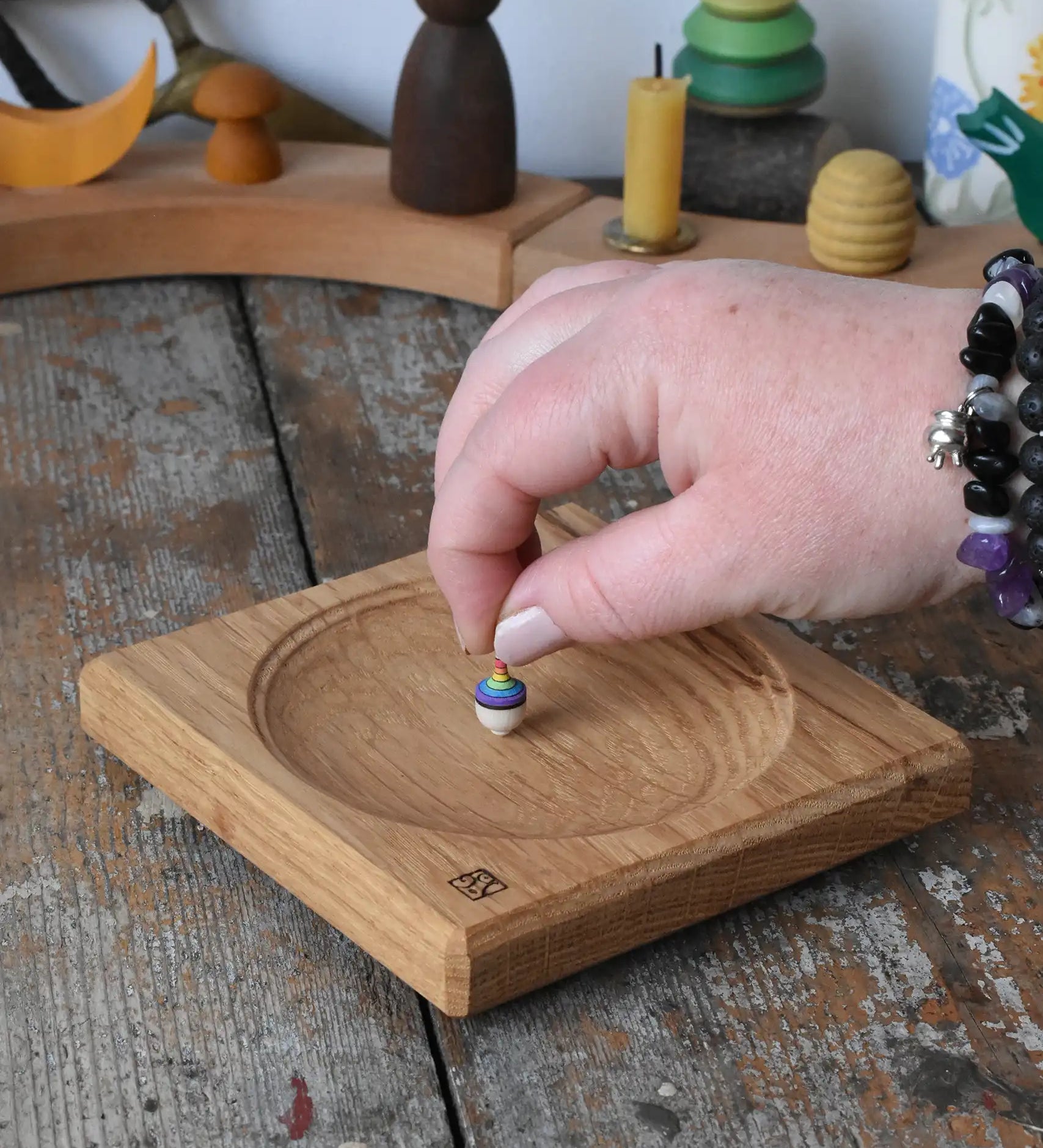 Mader Small Oak Spinning Plate placed on a wooden surface in the Babipur playroom. An adult's hand is placing a miniature rainbow spinning top on the board.