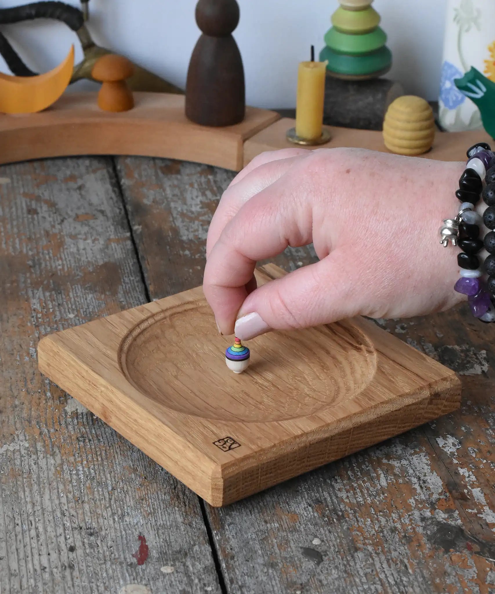 Mader Small Oak Spinning Plate placed on a wooden surface in the Babipur playroom. An adult's hand is placing a miniature rainbow spinning top on the board.