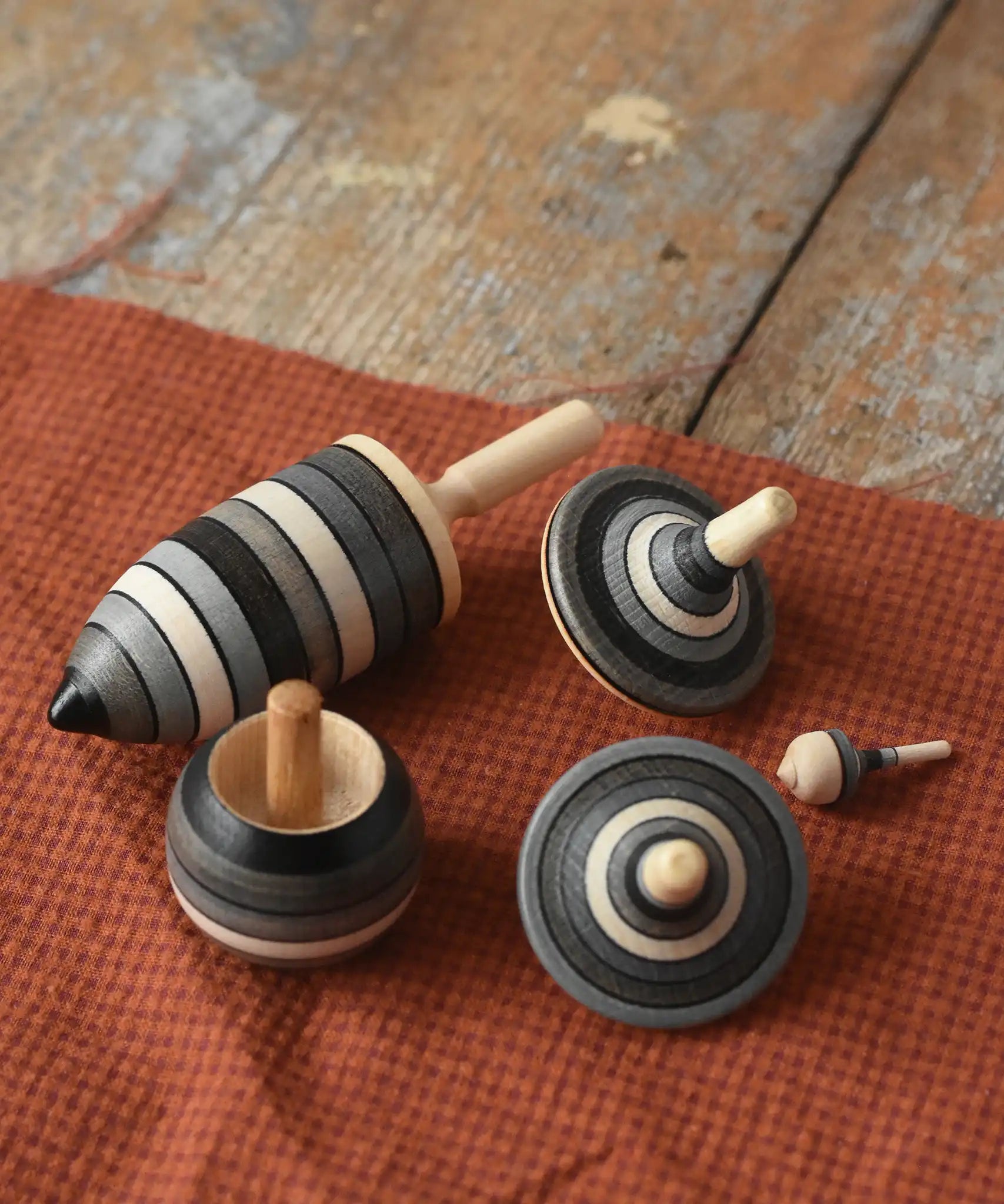Mader Kreiselmanufaktur graphite spinning top learning set placed on a cloth on a wooden surface in the Babipur playroom. The set has 5 striped tops in shades of grey, white and black.