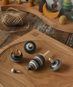 Mader Kreiselmanufaktur graphite spinning top learning set placed on an oak spinning plate on a wooden surface in the Babipur playroom. 