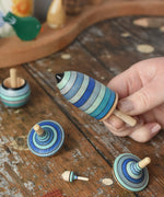 Mader Kreiselmanufaktur ice blue spinning top learning set on a wooden surface in the Babipur playroom. Close up of the thunderbolt spinner in an adult's hand.