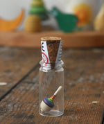 The miniature spinning top from the Mader Kreiselmanufaktur rainbow spinning top learning set inside it's glass, cork top jar placed on a wooden surface in the Babipur playroom. 
