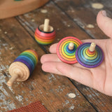 Mader Kreiselmanufaktur rainbow spinning top learning set placed on a wooden surface in the Babipur playroom. Two rainbow striped traditional tops are shown in an adult's hand.