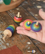 Mader Kreiselmanufaktur rainbow spinning top learning set placed on a wooden surface in the Babipur playroom. Two rainbow striped traditional tops are shown in an adult's hand.