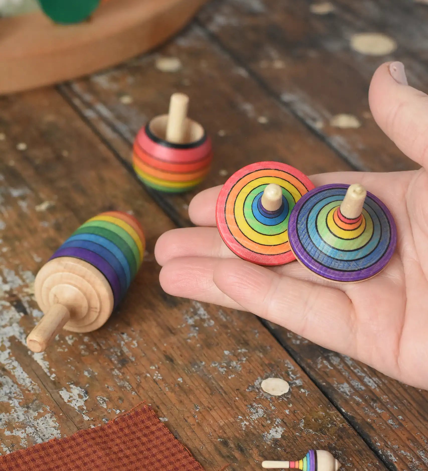 Mader Kreiselmanufaktur rainbow spinning top learning set placed on a wooden surface in the Babipur playroom. Two rainbow striped traditional tops are shown in an adult's hand.