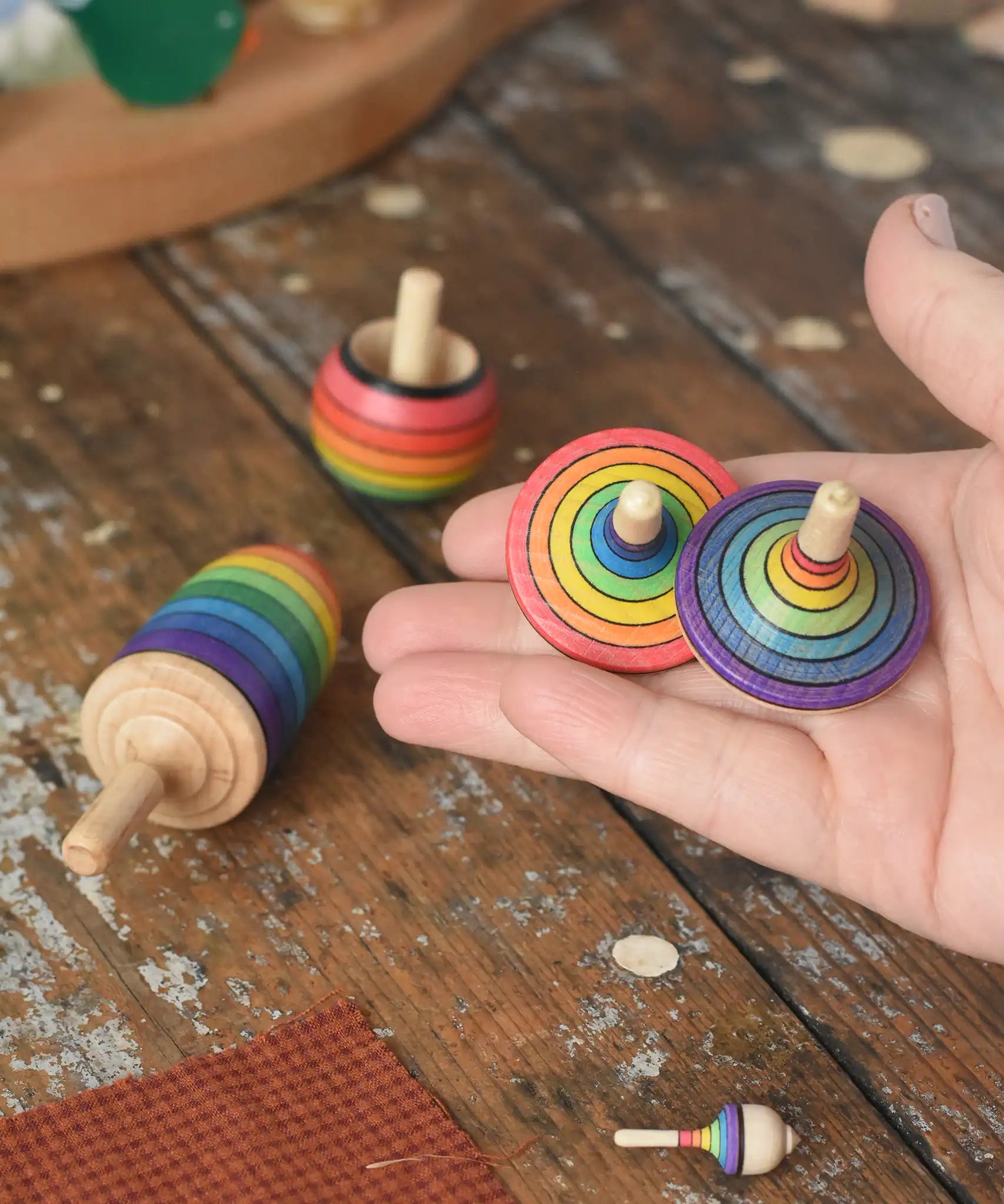 Mader Kreiselmanufaktur rainbow spinning top learning set placed on a wooden surface in the Babipur playroom. Two rainbow striped traditional tops are shown in an adult's hand.