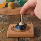 Mader Kreiselmanufaktur superstar wooden spinning top toy with and spinning plate placed on a wooden desk in the Babipur playroom. An adult's hand can be seen holding the spinner upright on the board. 