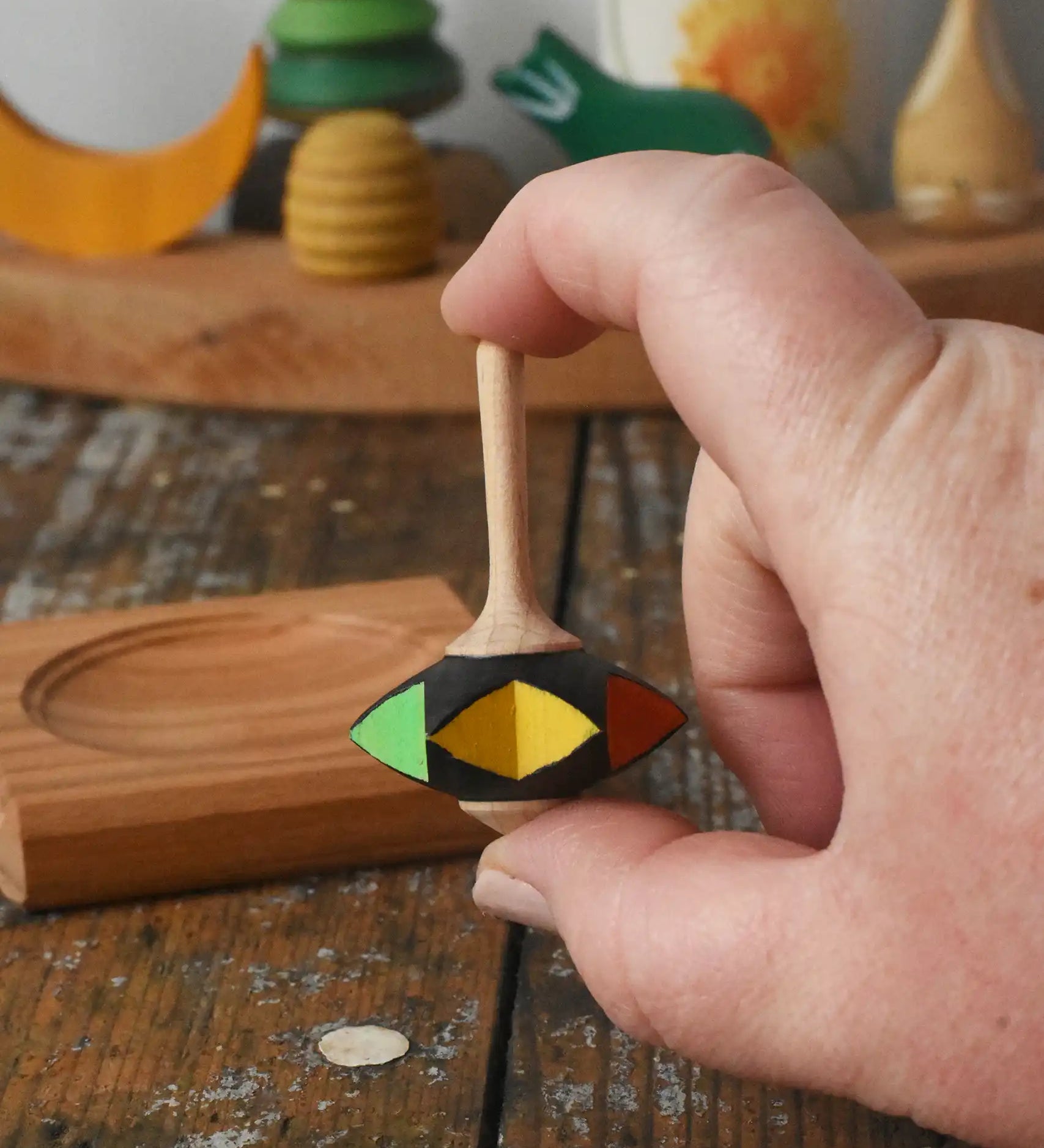 Mader Kreiselmanufaktur superstar wooden spinning top toy shown in an adult's hand with and spinning plate placed on a wooden desk in the Babipur playroom.