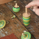 Mader Kreiselmanufaktur green grass spinning top learning set on a wooden surface in the Babipur playroom. An adult's hand is placed on the thunderbolt spinner.