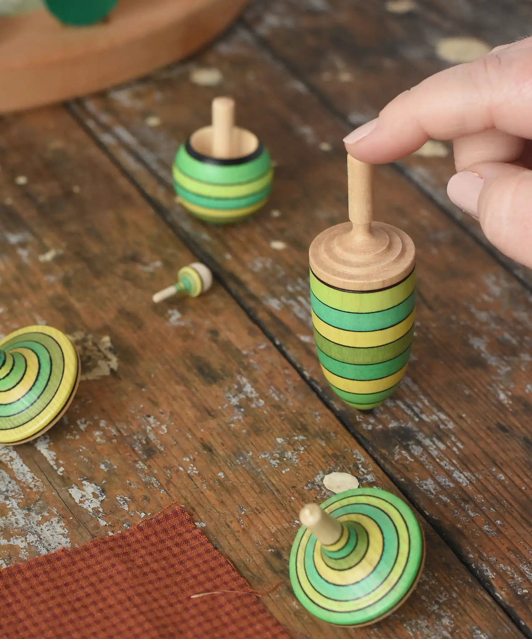 Mader Kreiselmanufaktur green grass spinning top learning set on a wooden surface in the Babipur playroom. An adult's hand is placed on the thunderbolt spinner.