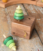 Mader Kreiselmanufaktur forest music box placed on a wooden desk in the Babipur playroom. A wooden wind up musical box with two slots for the green tree shaped spinners on the top. One tree has been taken out of the box.