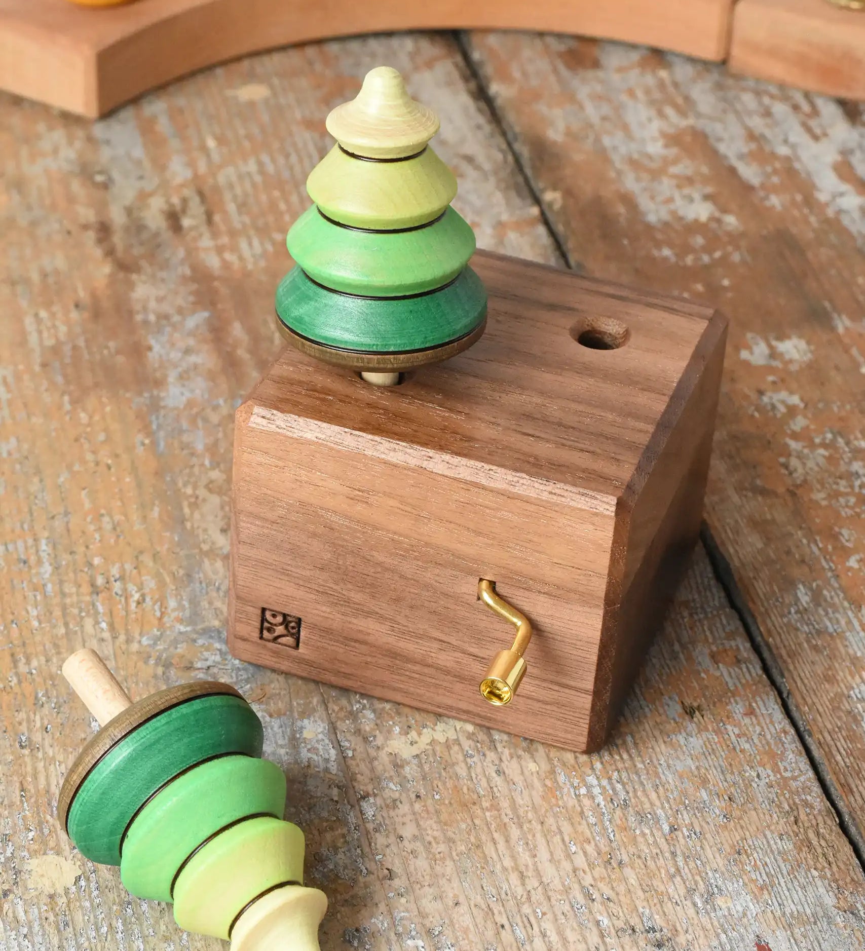 Mader Kreiselmanufaktur forest music box placed on a wooden desk in the Babipur playroom. A wooden wind up musical box with two slots for the green tree shaped spinners on the top. One tree has been taken out of the box.
