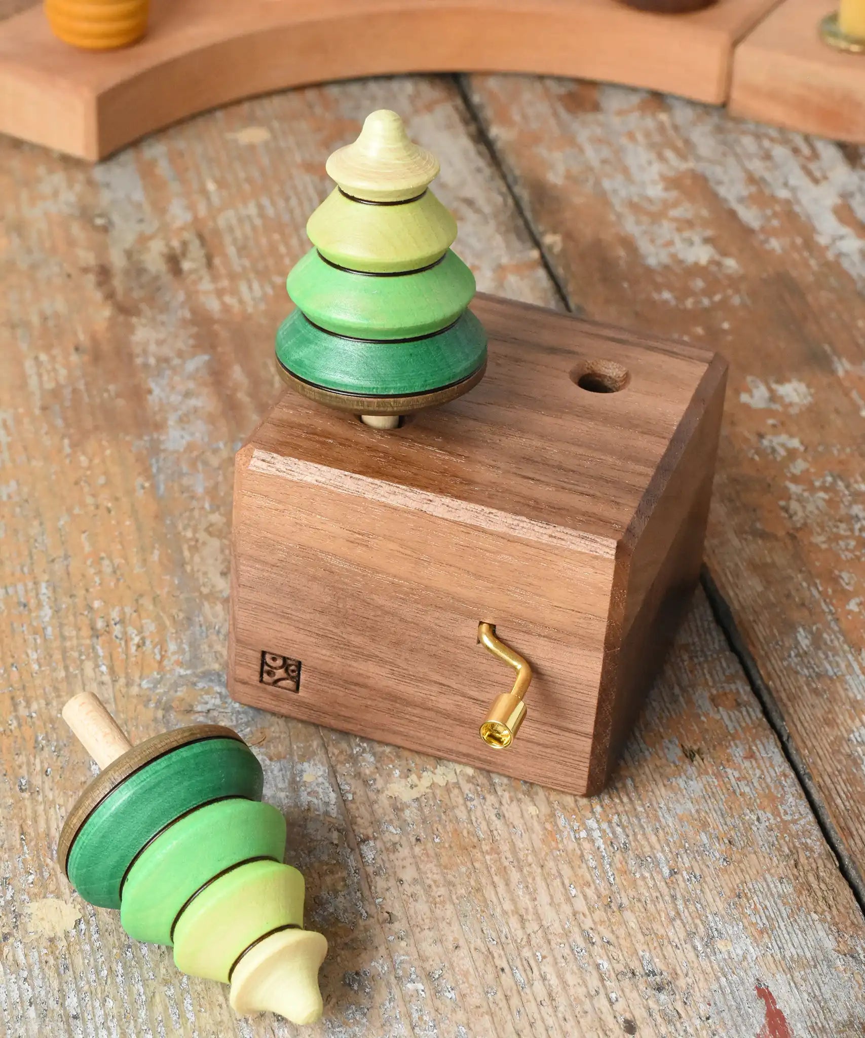 Mader Kreiselmanufaktur forest music box placed on a wooden desk in the Babipur playroom. A wooden wind up musical box with two slots for the green tree shaped spinners on the top. One tree has been taken out of the box.