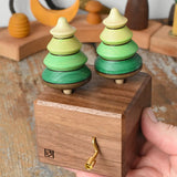 Mader Kreiselmanufaktur forest music box placed in an adult's hand. A wooden wind up musical box with two slots for the green tree shaped spinners on the top.