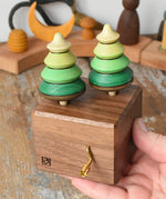 Mader Kreiselmanufaktur forest music box placed in an adult's hand. A wooden wind up musical box with two slots for the green tree shaped spinners on the top.