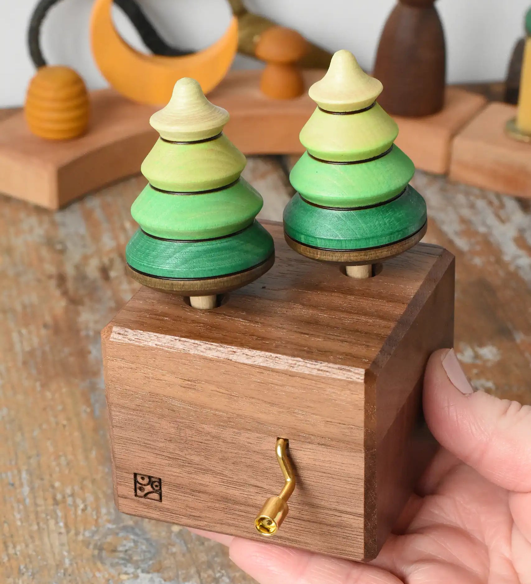 Mader Kreiselmanufaktur forest music box placed in an adult's hand. A wooden wind up musical box with two slots for the green tree shaped spinners on the top.