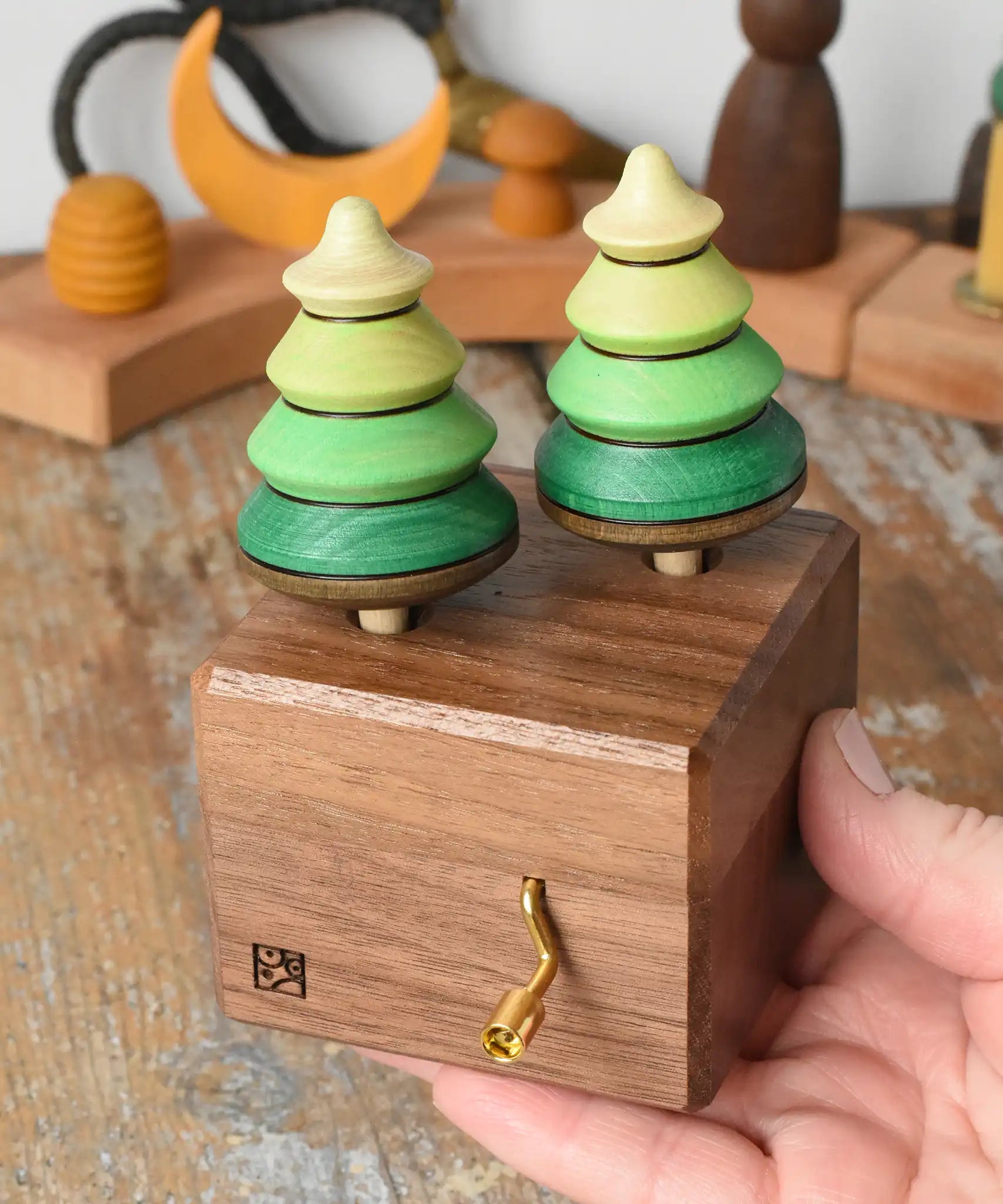 Mader Kreiselmanufaktur forest music box placed in an adult's hand. A wooden wind up musical box with two slots for the green tree shaped spinners on the top.