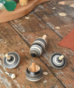 Mader Kreiselmanufaktur graphite spinning top learning set placed on a wooden surface in the Babipur playroom. The set has 5 striped tops in shades of grey, white and black.