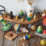 Mader traditional handmade wooden spinning top toys paced on a wooden desk in the Babipur playroom. Green my first spinning top with starter, ring spinning top, small helene's captive rings on plate, rainbow thunderbolt, rainbow turn-over, rainbow mini, bee, and forest set.