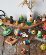 Mader traditional handmade wooden spinning top toys paced on a wooden desk in the Babipur playroom. Green my first spinning top with starter, ring spinning top, small helene's captive rings on plate, rainbow thunderbolt, rainbow turn-over, rainbow mini, bee, and forest set.