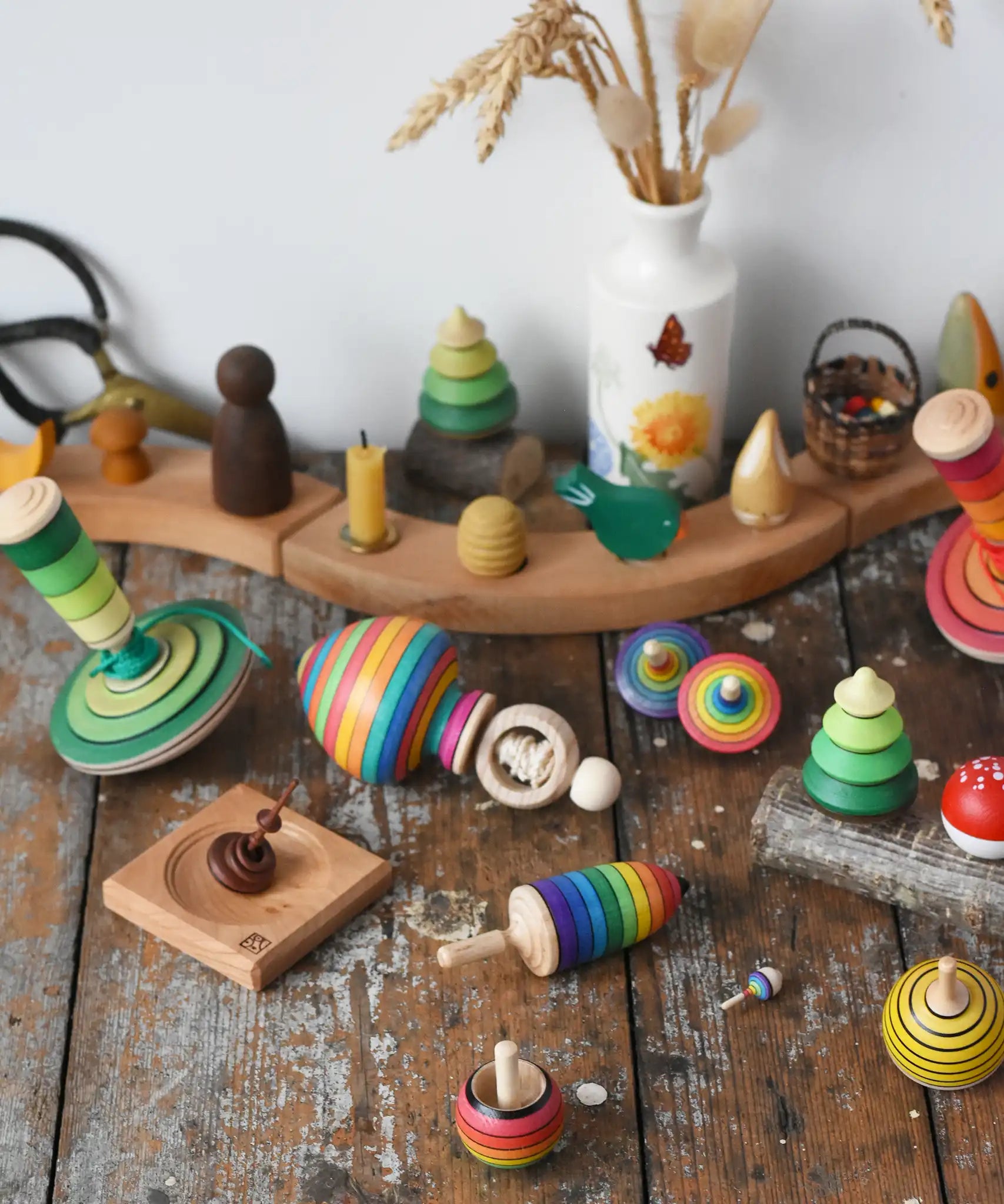 Mader traditional handmade wooden spinning top toys paced on a wooden desk in the Babipur playroom. Green my first spinning top with starter, ring spinning top, small helene's captive rings on plate, rainbow thunderbolt, rainbow turn-over, rainbow mini, bee, and forest set.