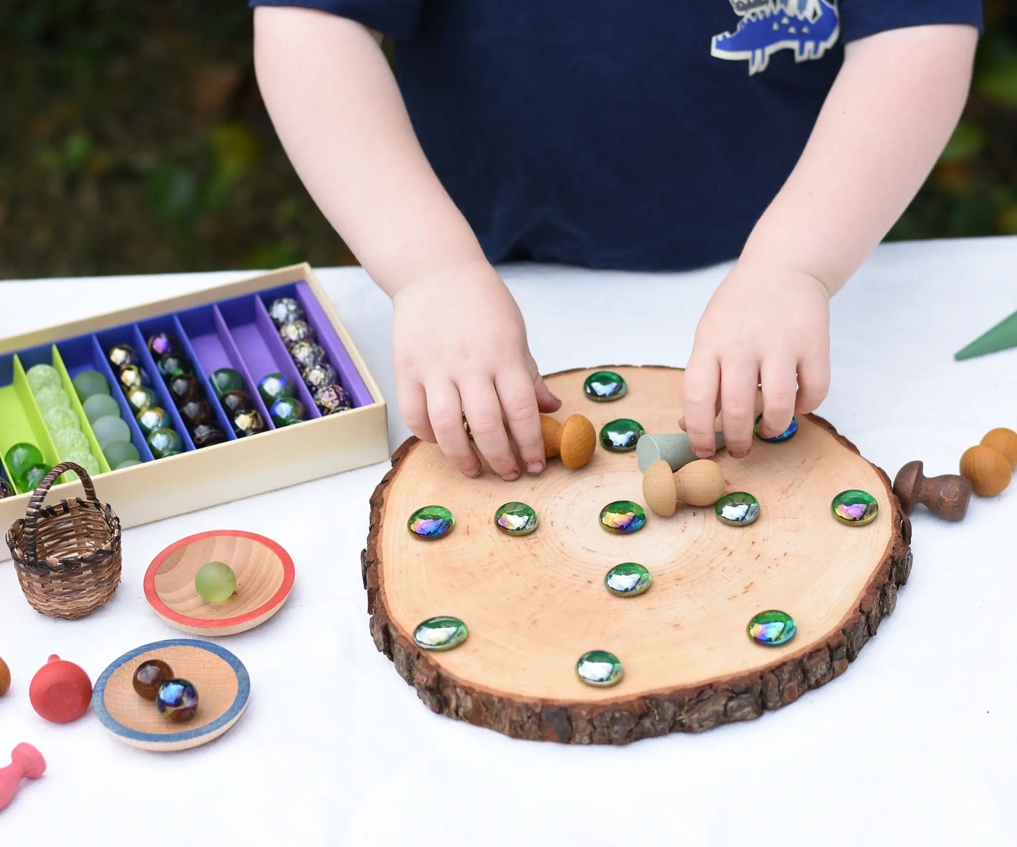 Child placing marbles and mandala pieces on a log slice from the Magic Wood Eco Blocks Big Box 70 piece set, decorating an imaginary pizza.
