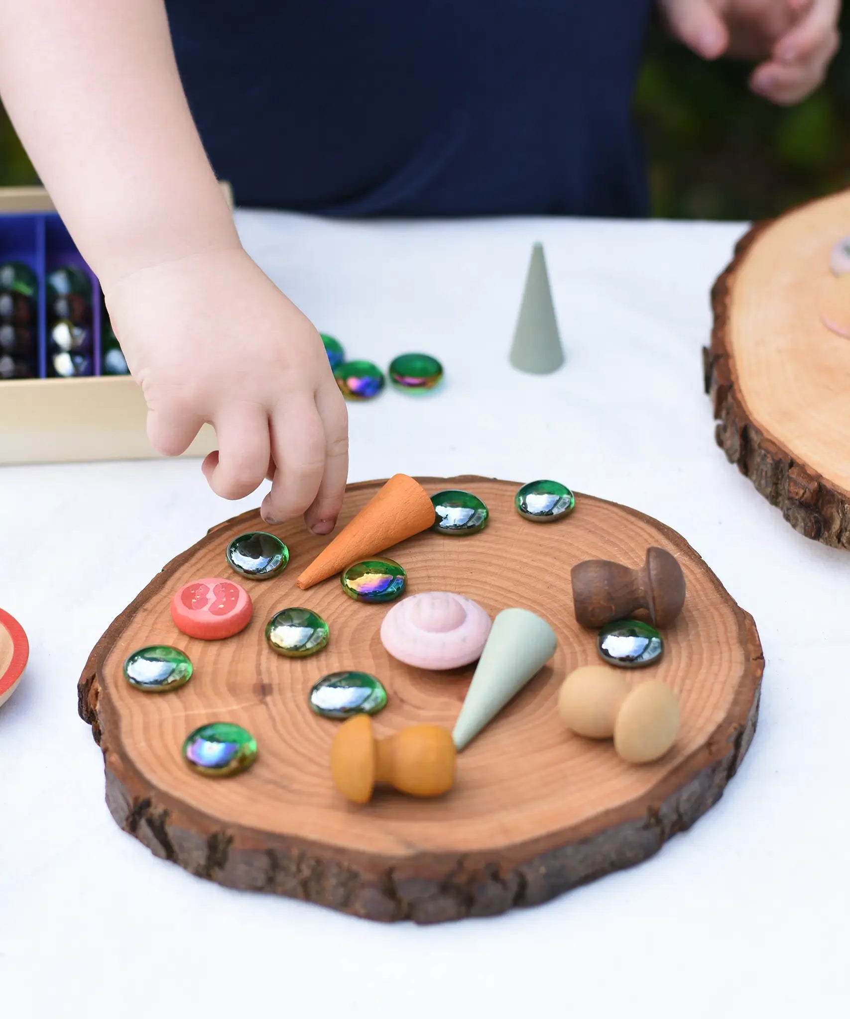 Child placing flat glass marbles and Grapat mandala pieces on a large log slice from the Magic Wood Eco Blocks Big Box 70 piece set.