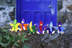 The Magic Wood Six Felt Elves shown lined up in seated positions on a stone wall ourtdoors.