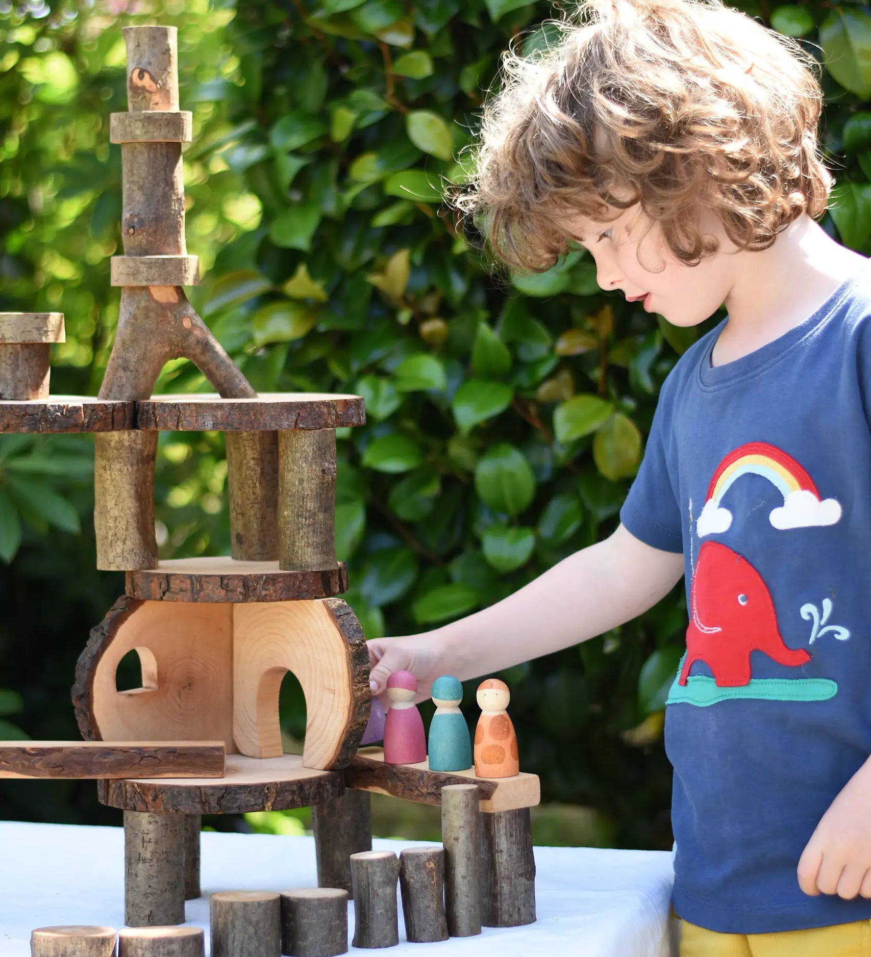 Magic Wood oiled ecoBlocks in a 36 piece set used to build a structure with a child wearing a Babipur t-shirt placing wooden peg dolls on it. 