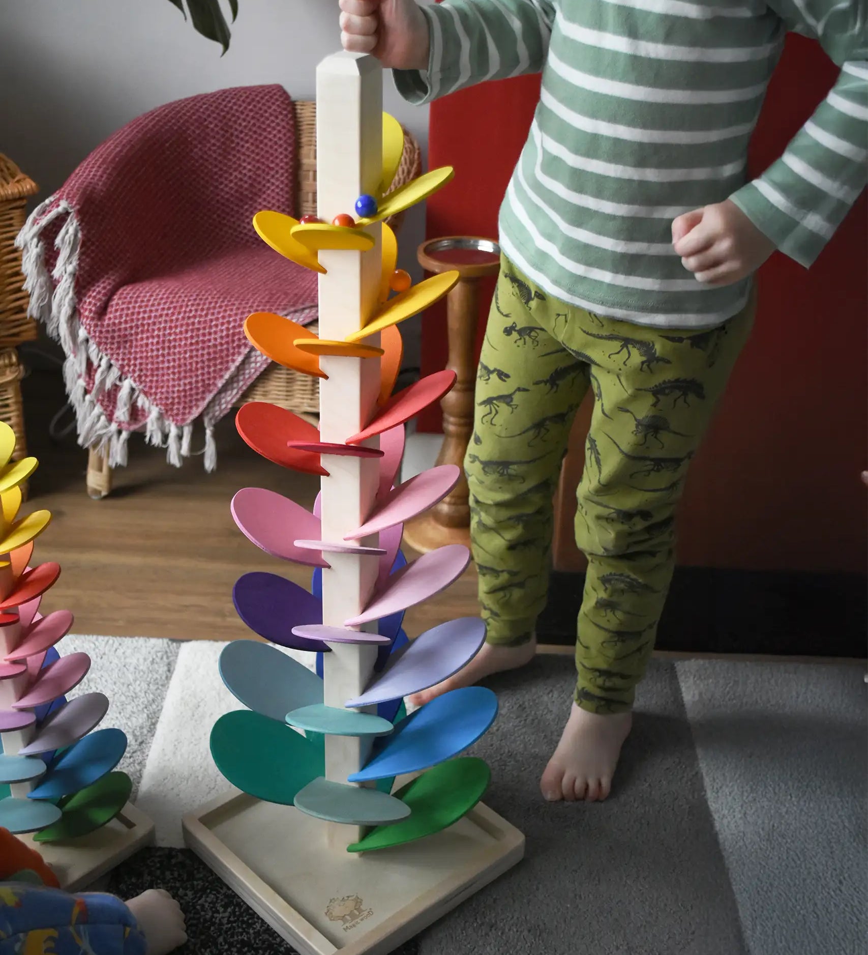 A close up of a child standing next to a Magic Wood 72cm large marble tree placing glass marbles on the top leaves. Marbles are cascading down the rainbow coloured leaves.