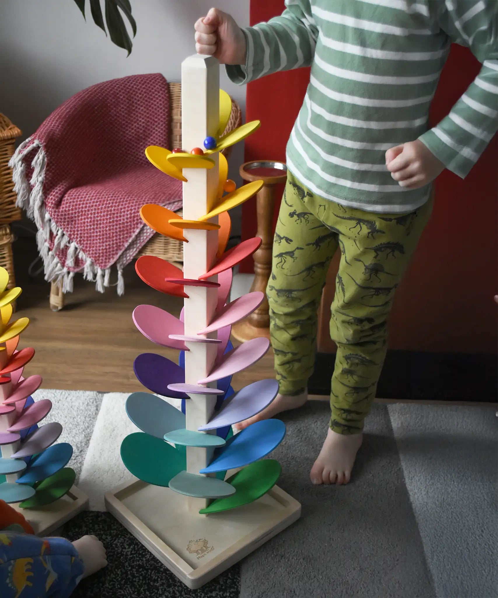 A close up of a child standing next to a Magic Wood 72cm large marble tree placing glass marbles on the top leaves. Marbles are cascading down the rainbow coloured leaves.