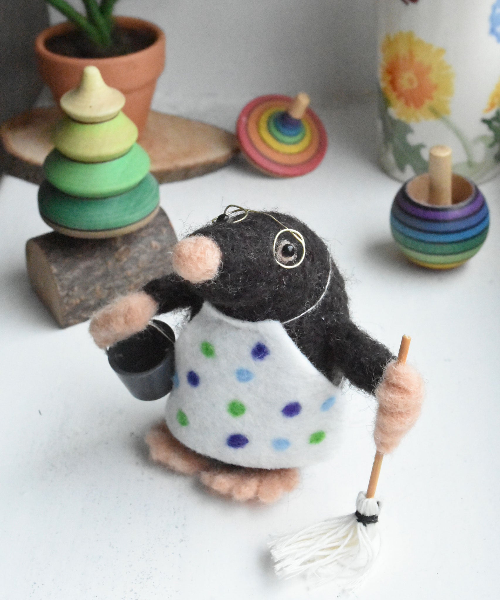 A felted mole figure wearing a spotty apron holding a bucket and a mop. The figure has been created using The Makerss Small Busy Mr Mole Needle Felt Kit.