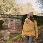 A woman wearing the Mamalila Allweather Cosy Allrounder Babywearing Jacket in a Mustard yellow colour carrying a baby using the babywearing insert.